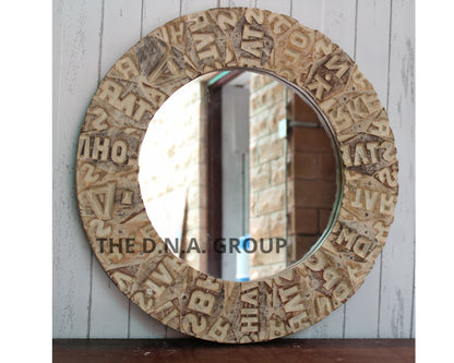 Round mirror with decorative frame featuring letters, on a wooden surface with a white wall background.